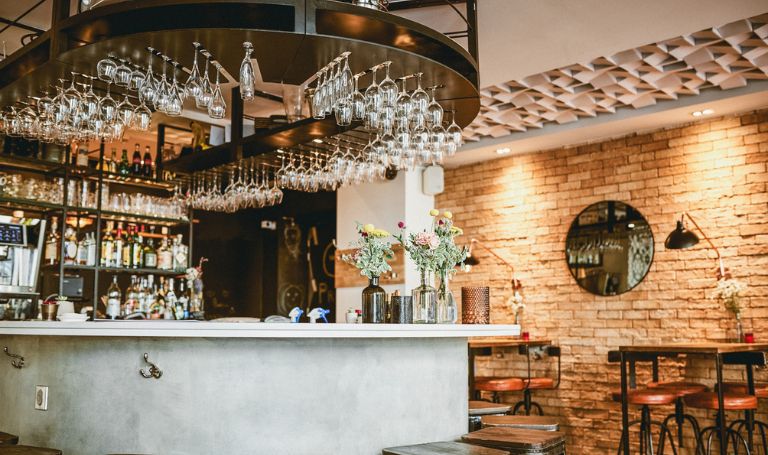 En modern bar med svängd disk, hängande vinglas, blomvaser, exponerade tegelväggar, en rund spegel och höga trästolar designade med precision som finns i arkitektritningar. Baren är väl upplyst och ståtar med olika flaskor på hyllorna.